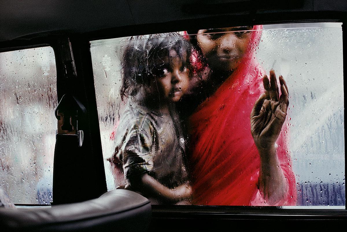 Mutter und Kind am Autofenster, Bombay von Steve McCurry ist ein 20 x 24 Zoll großer digitaler C-Print, erhältlich in einer Auflage von 90 Stück. Dieses Foto zeigt eine Frau in Rot, die ein Kind hält und durch ein verregnetes Fenster in ein Auto