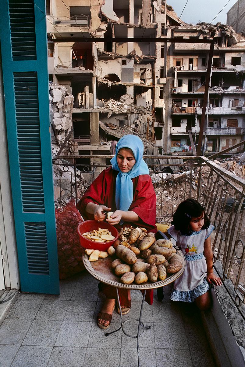 Mutter und Tochter auf ihrem Balkon in Beruit von Steve McCurry zeigt eine Frau und ein Kind, die im Freien sitzen. Die Frau blickt nach unten, konzentriert auf das Schälen ihrer Kartoffeln, während das Mädchen neben ihr kniet und hinter dem