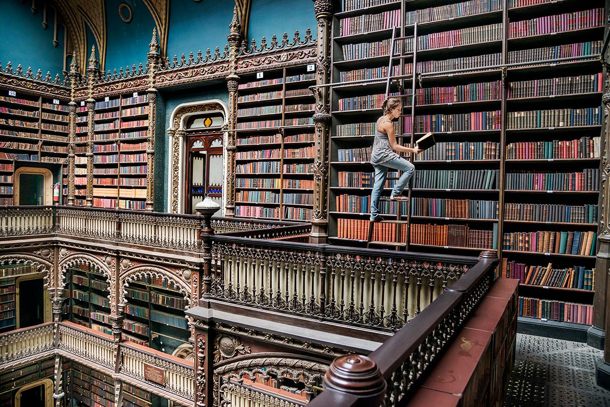 Real Gabinete Português de Leitura, which translates to the Royal Portuguese Cabinet of Reading, by Steve McCurry depicts a woman standing on a ladder in an extravagant library. She pauses to read a book, the ladder resting against the towering
