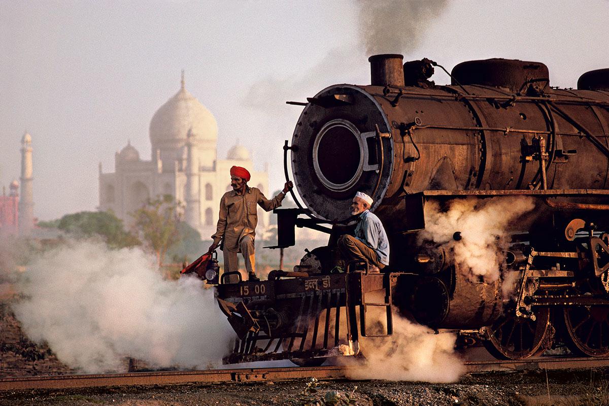 Taj and Train by Steve McCurry is a 20 x 24 inch digital c-print on FujiFlex Crystal Archive Supergloss paper, available in an edition of 90. This photograph features two men holding on to the front of a train, while the train is passing in front of