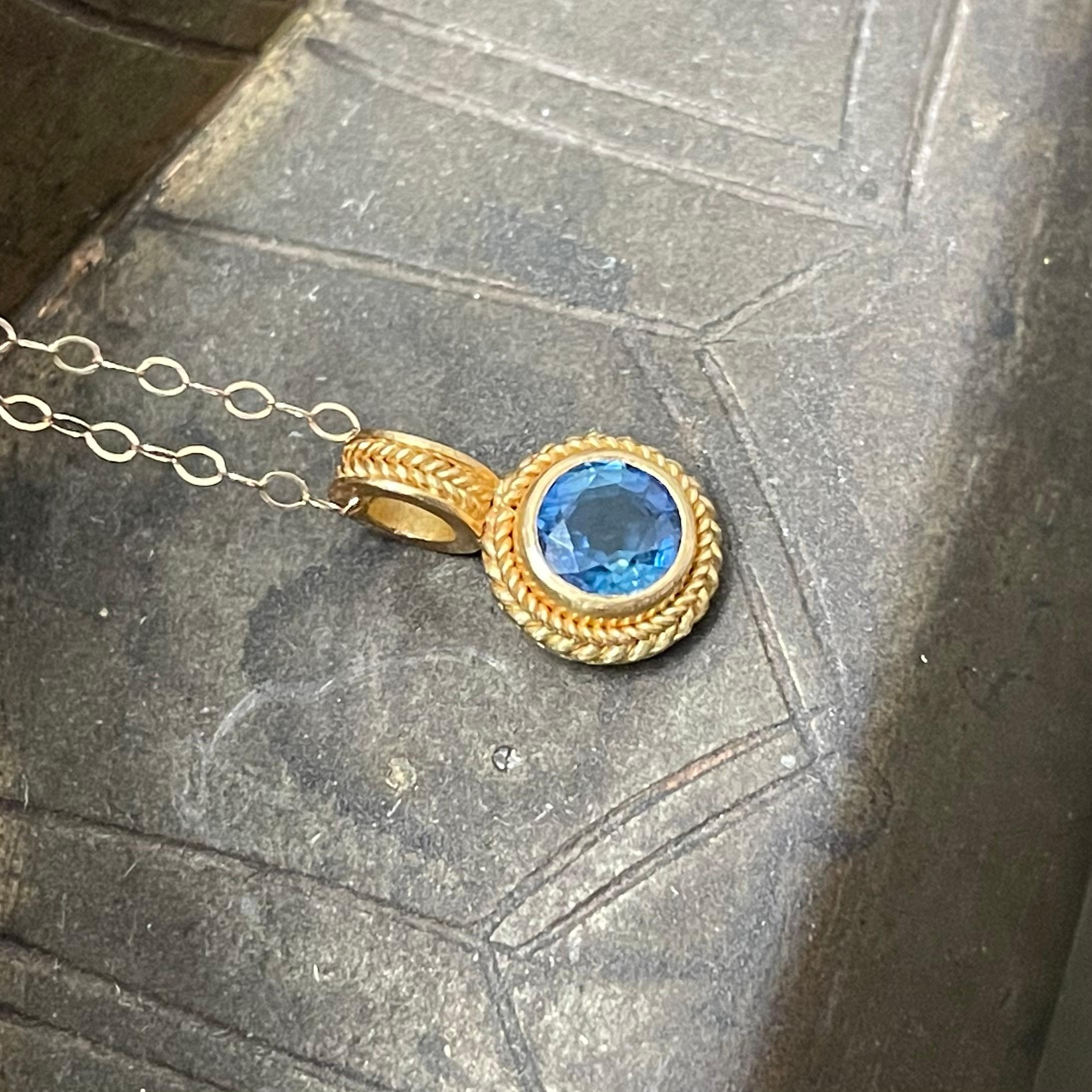 A light blue 5mm round faceted blue sapphire is set in a 22K double twist wire bezel setting with a round loop above decorated with similar motif.  This pendant comes with a delicate 18 inch 14K gold cable chain (could also be nice with a heavier