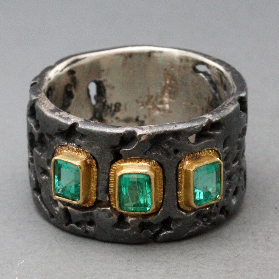 Three crystalline light green 3 x 4 mm octagonal cut Columbian emeralds are surrounded by lined 18K gold bezels and set within an organically textured wide darkly oxidized cigar band shank to create this unique sharply contrasting design.  This ring