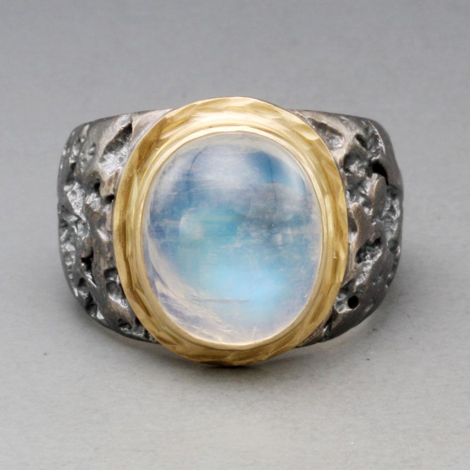 A luminous 12 x 15 mm oval rainbow moonstone rests atop an organically textured wide oxidized sterling silver band surrounded by a hammered 18K bezel accent currently in this beautifully contrasting creation by Steven Battelle.  Currently sized 8.