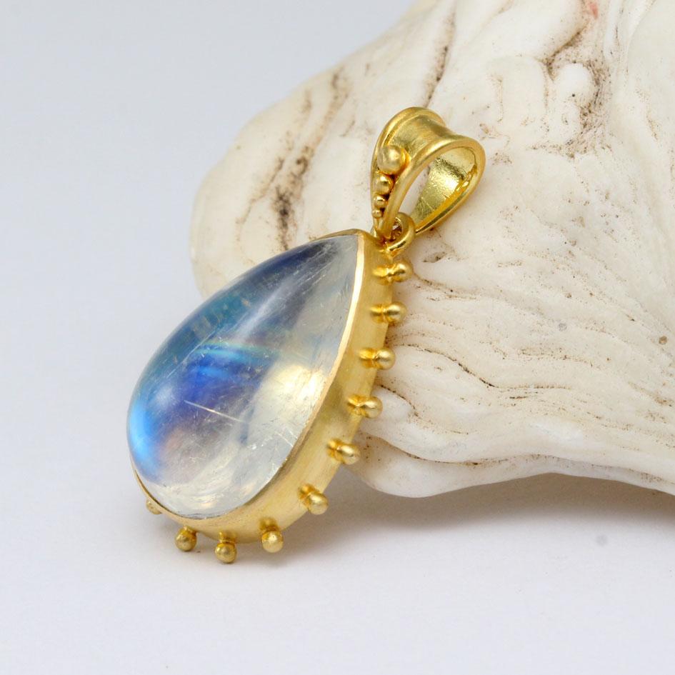 A lively 10 x 17 mm pear shaped rainbow moonstone cabochon shimmers with blue and gold highlights within a simple matte-finish 18K gold bezel surrounded by spaced 