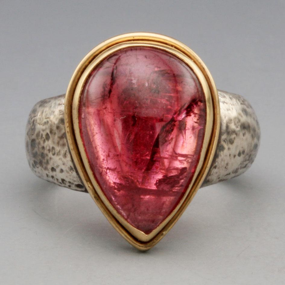 A lively and vibrant 12 x 17 mm pear shaped Mozambique pink tourmaline cabochon is held in a simple 18K gold bezel with additional 18k gold wire double bezel below, all atop a wide and tapered sterling silver shank textured with a hammered and