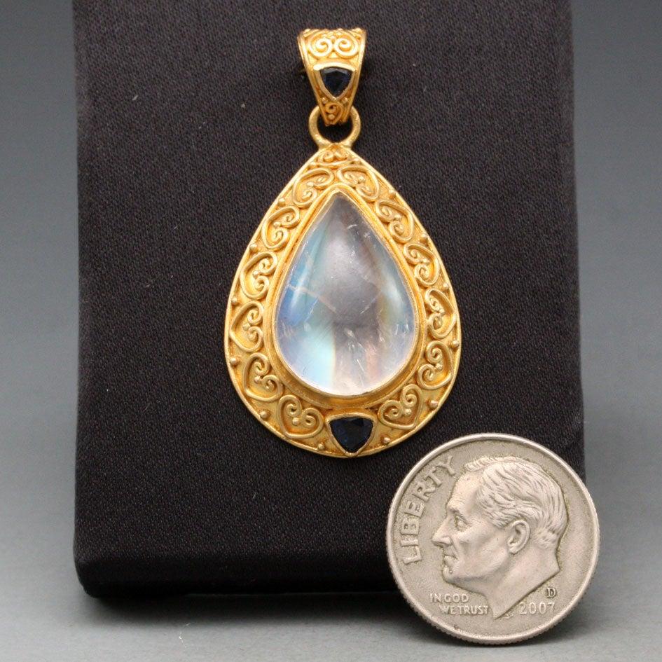 A magical 12x18mm pear shaped rainbow moonstone cabochon displaying a wide range of colors is accented with two trillium faceted blue sapphires totaling 0.8 carats set in this hand fabricated fine granulation and wire work ('bun