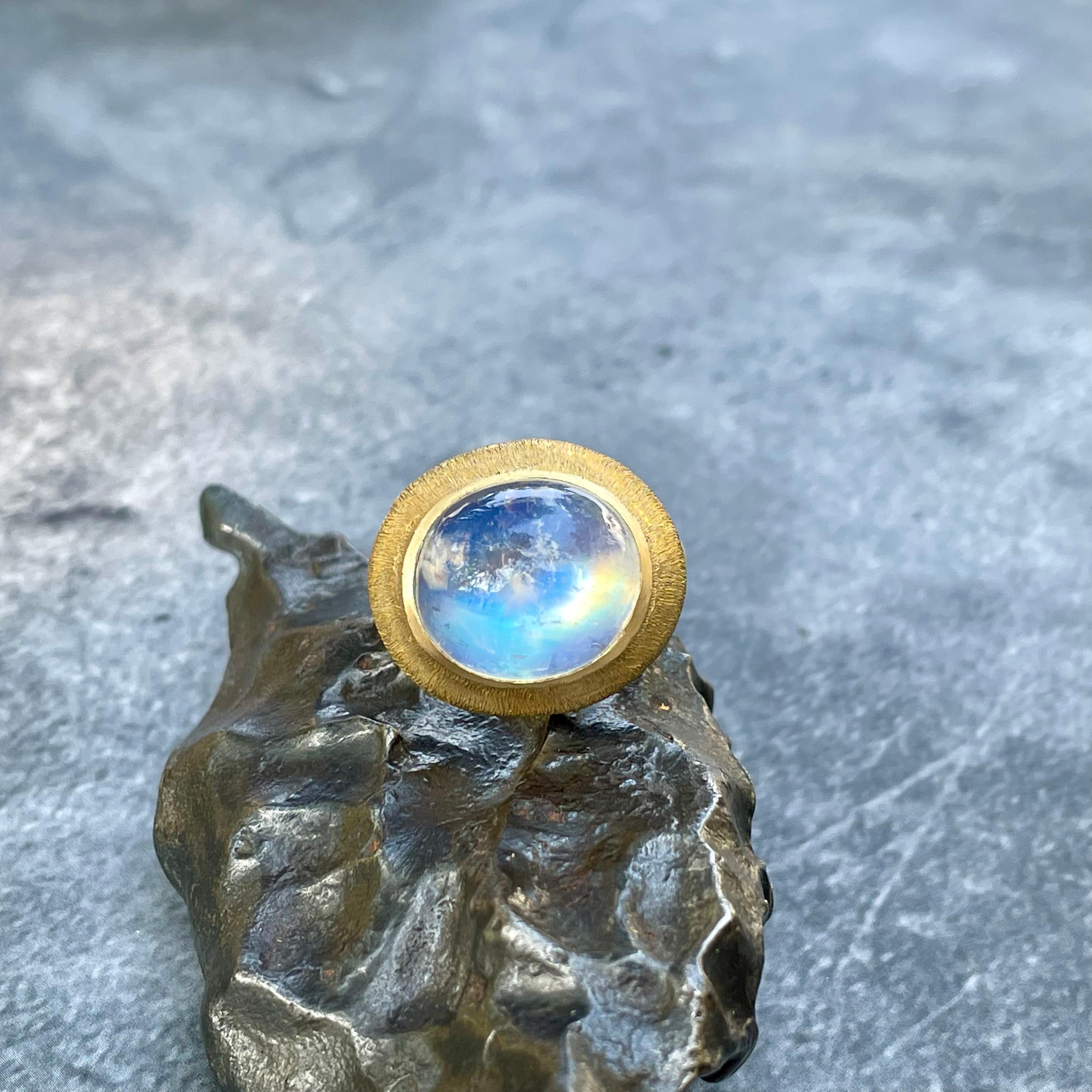 A magical high dome 12 x 14 mm rainbow moonstone cabochon enchants surrounded by an organic line texture wide sculpted bezel in this classically simple 18 K ring design. The muted texture of the bezel is a beautiful complement to the shimmering