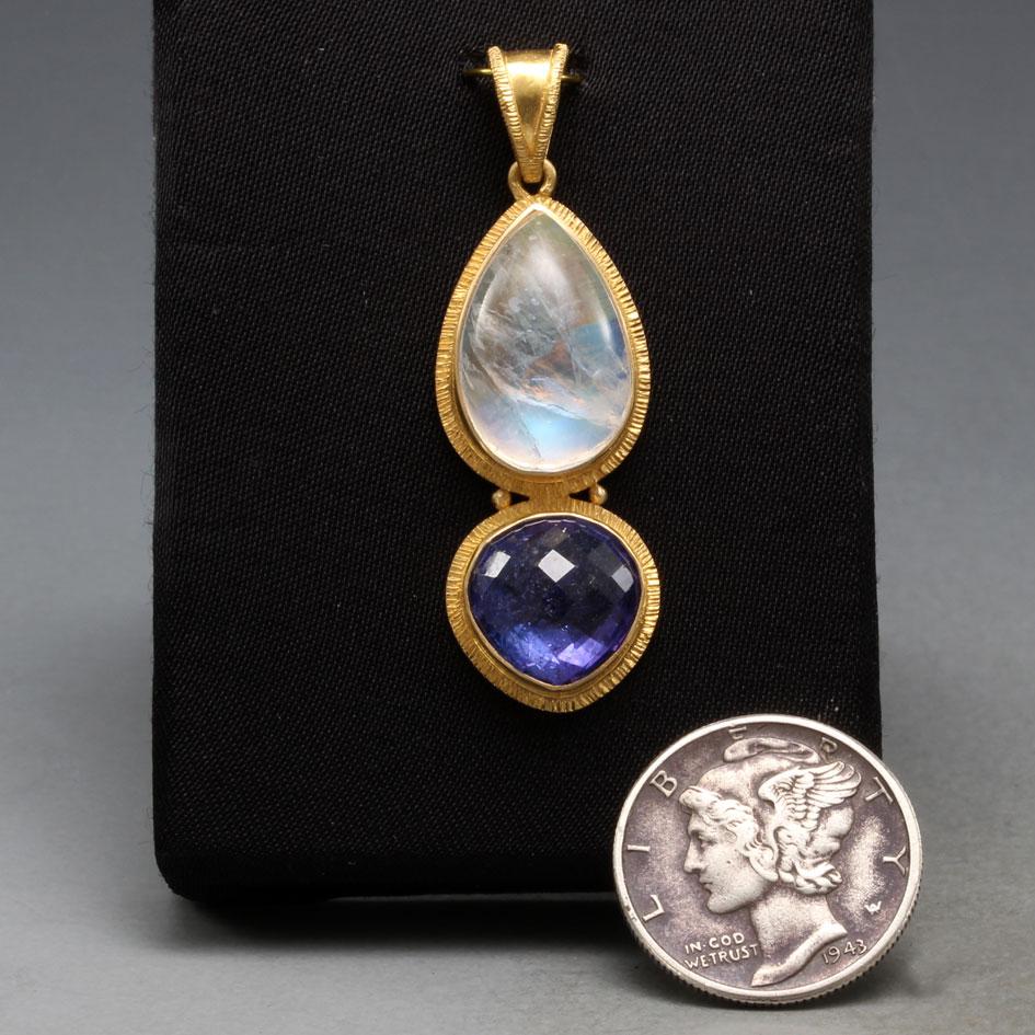 An 11 mm trillium rose faceted tanzanite rests below a 10x15 mm pear shaped rainbow moonstone cabochon, all surrounded by line textured 18K gold double bezels and with 2 