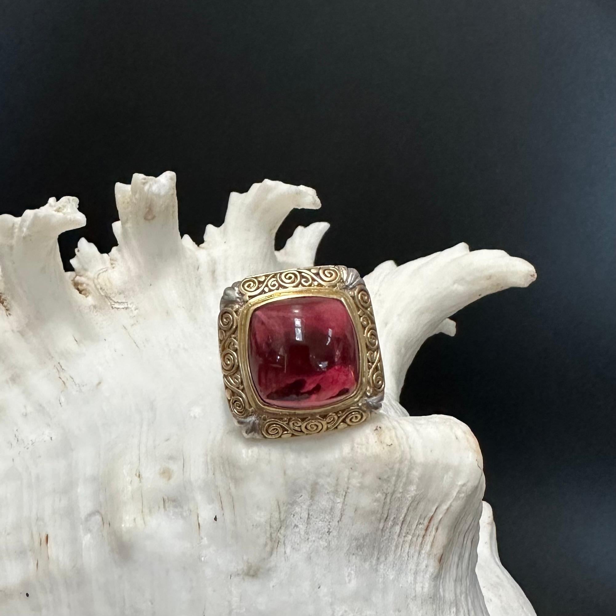 A lively and clear 16 mm cushion shaped cabochon pink tourmaline is set in an 18K gold bezel surrounded by intricate Byzantine style artisanal 