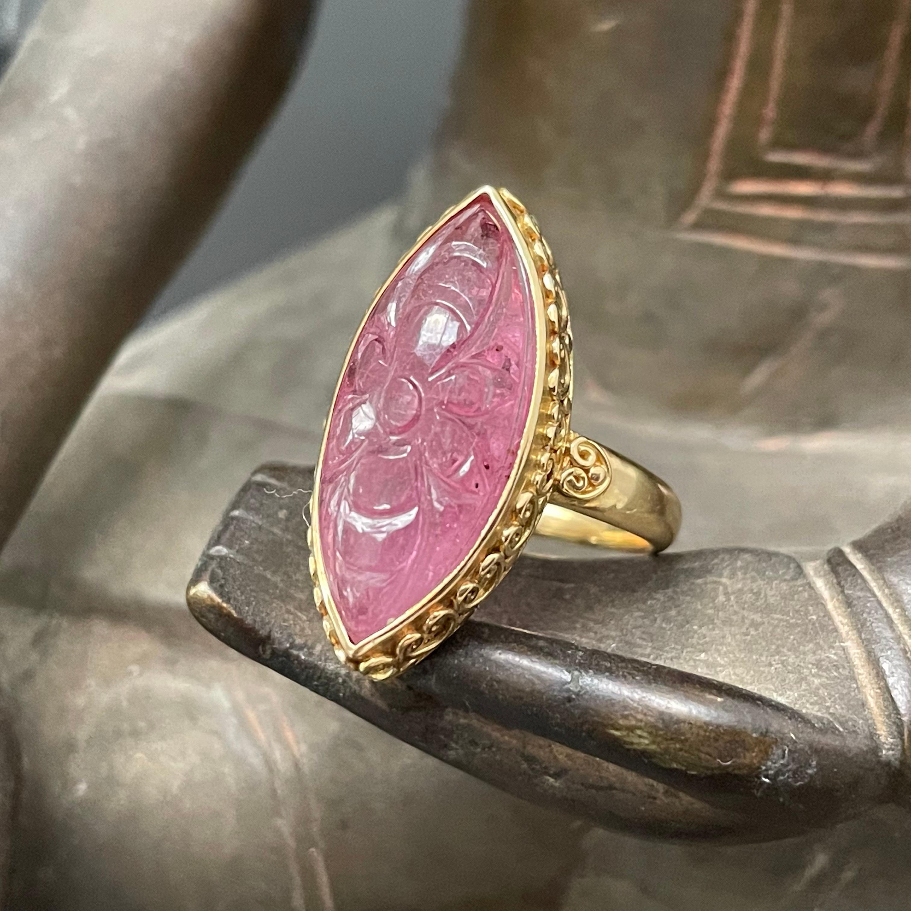 A 10 x 22 mm marquis shaped flower carved pink ruby cabochon is held in a handmade 18K gold 