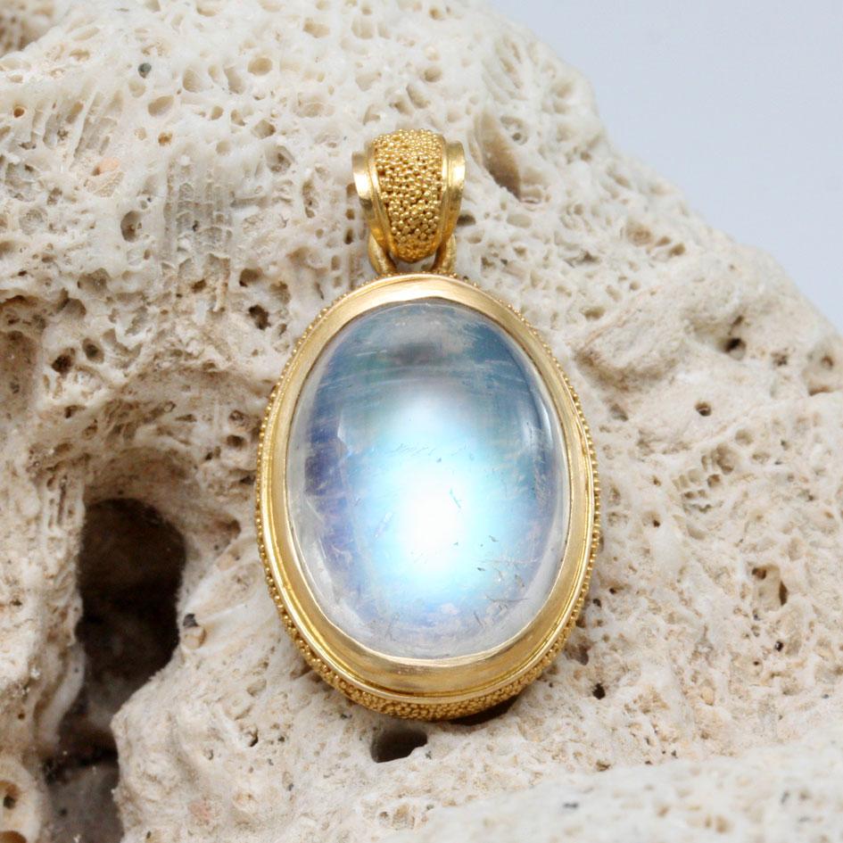 A luminous blue flashing 13 x 18 mm oval rainbow moonstone cabochon rests in a slightly cupped high-karat bezel surrounded by delicate 