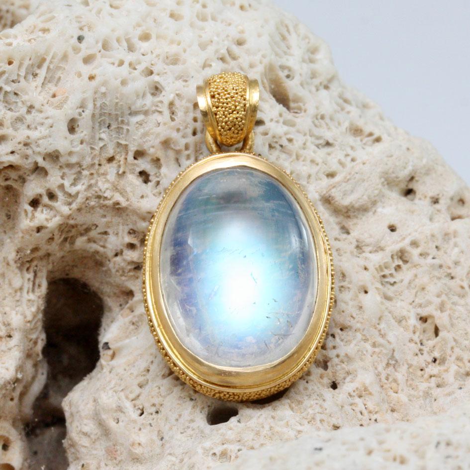 Ein leuchtend blau blinkender, 13 x 18 mm großer, ovaler Regenbogen-Mondstein-Cabochon ruht in einer leicht gewölbten, hochkarätigen Lünette, umgeben von einer zarten 