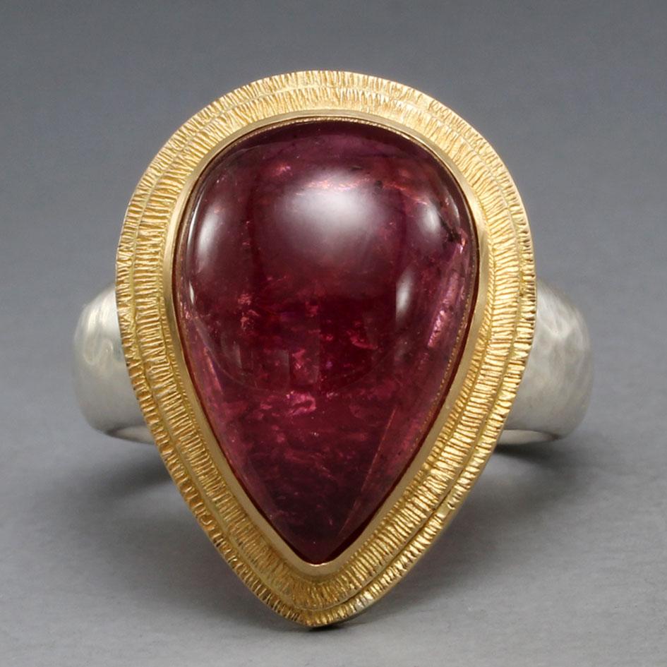 Un vivace e succoso cabochon di tormalina rosa a forma di pera di 12 x 17 mm è racchiuso in un castone artigianale in oro 18 carati circondato da due accenti a doppio castone a 