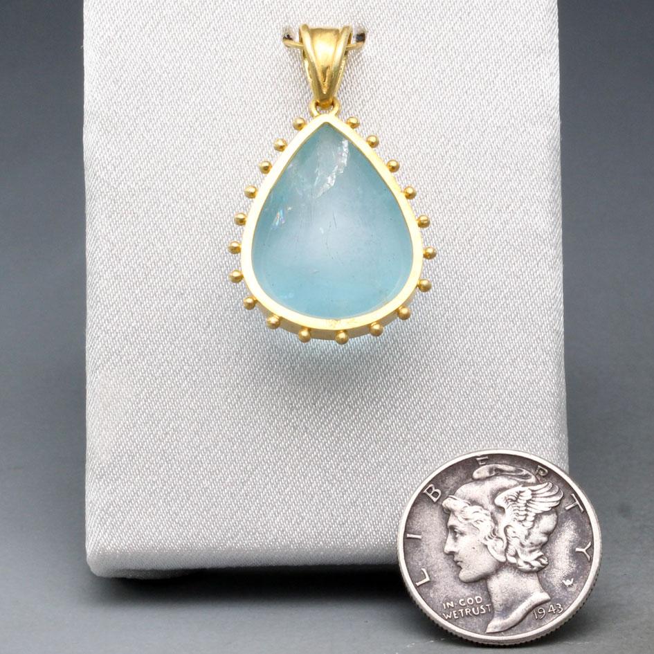 A limpid sea-blue 15 x 20 mm pear shaped cabochon aquamarine is held in a matte-finish 18K bezel surrounded by separated 