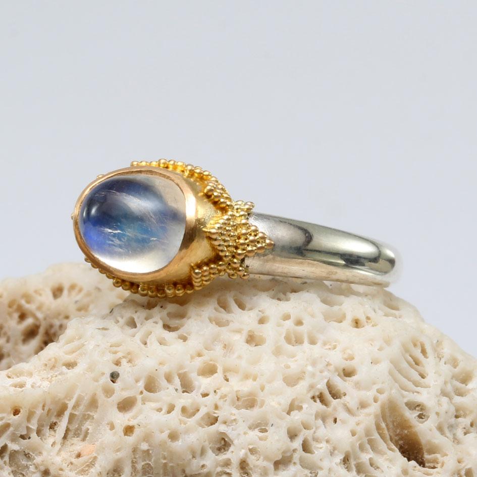 Un cabochon de pierre de lune arc-en-ciel bleue de 6 x 8 mm est serti dans de l'or fin riche et incrusté de petites granulations 