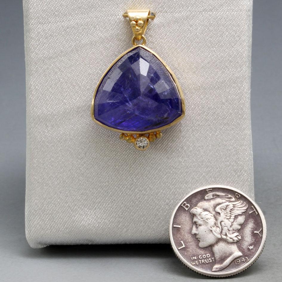 A deep blue 16 mm trillium faceted Tanzanite is held in a simple matte-finish 18K gold bezel with a 1..8 mm VS1 diamond below accented with graduated 