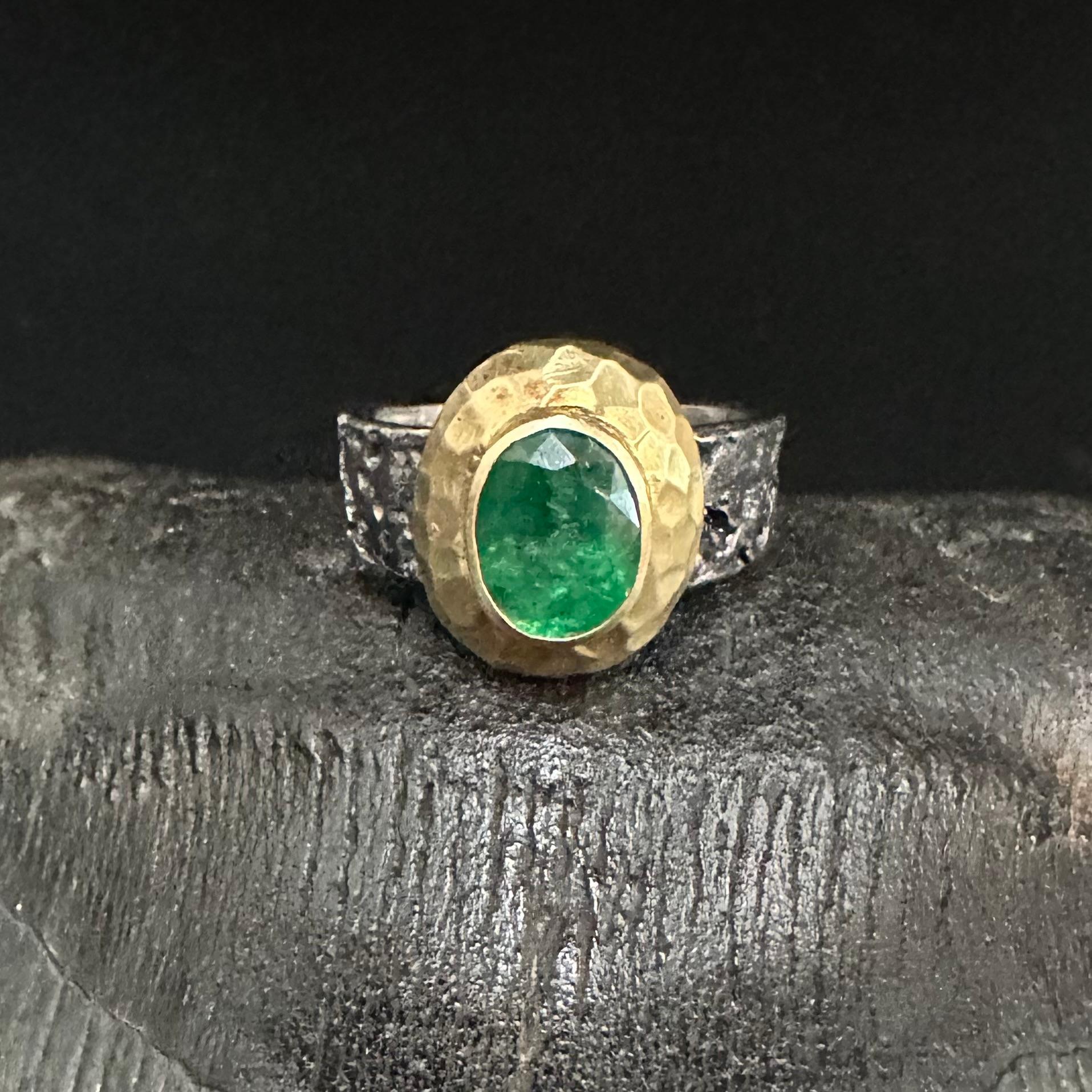 Ein lebendiger, ovaler, 7 x 9 mm großer, facettierter sambischer Smaragd ist in einer handgefertigten, breiten Lünette aus 18-karätigem Gold mit Hammerstruktur gefasst, die auf einem organisch strukturierten, tief oxidierten, breiten Sterling-Schaft