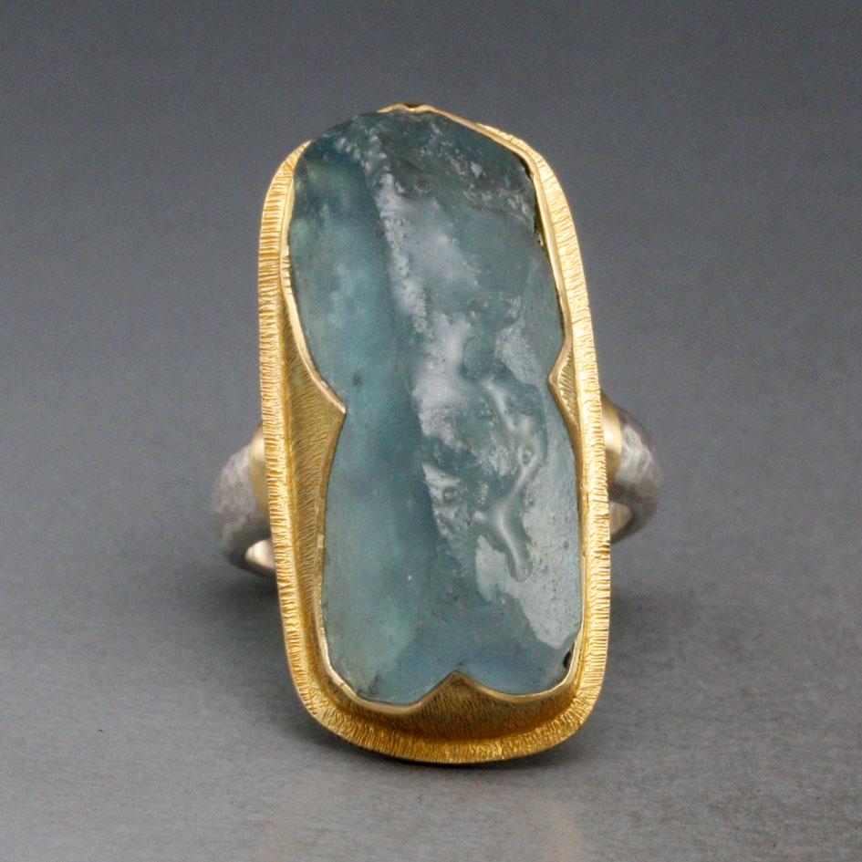 A slightly tumbled 11 x 26 mm roughly rectangular chunk of crystalline aquamarine is set within an ancient-inspired handmade 
