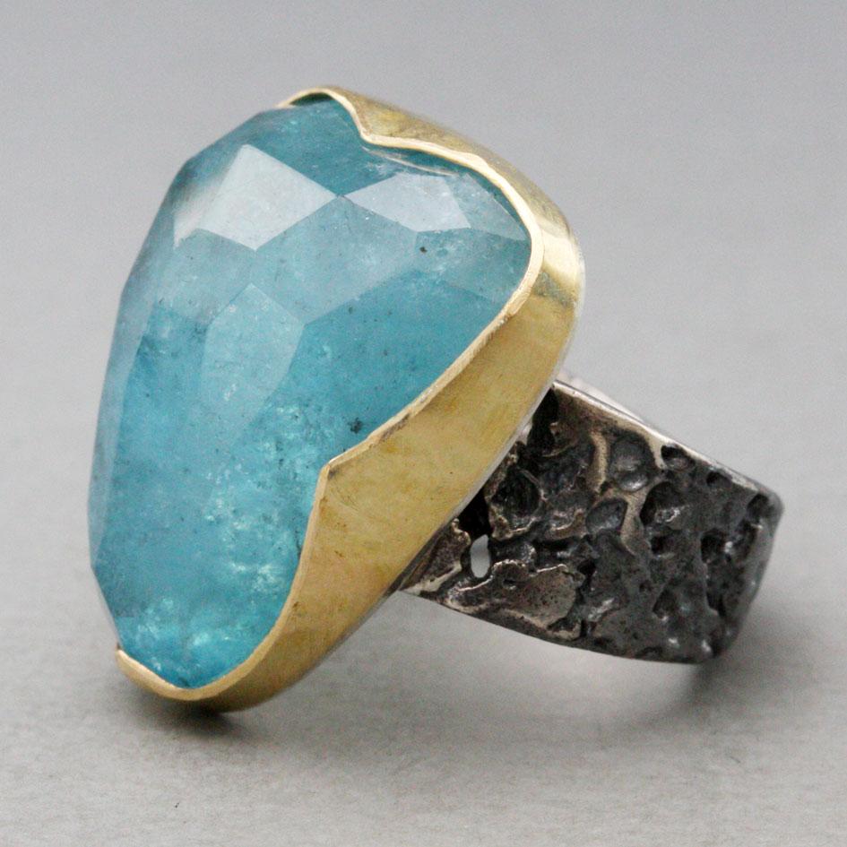 Ein schöner blauer, etwas dreieckiger und unregelmäßiger 18 x 22 mm großer Aquamarin im Rosenschliff mit Facettenschliff ist in diesem Design in eine gehämmerte und strukturierte, einfache Lünette aus 18 Karat Gold auf einem breiten oxidierten,