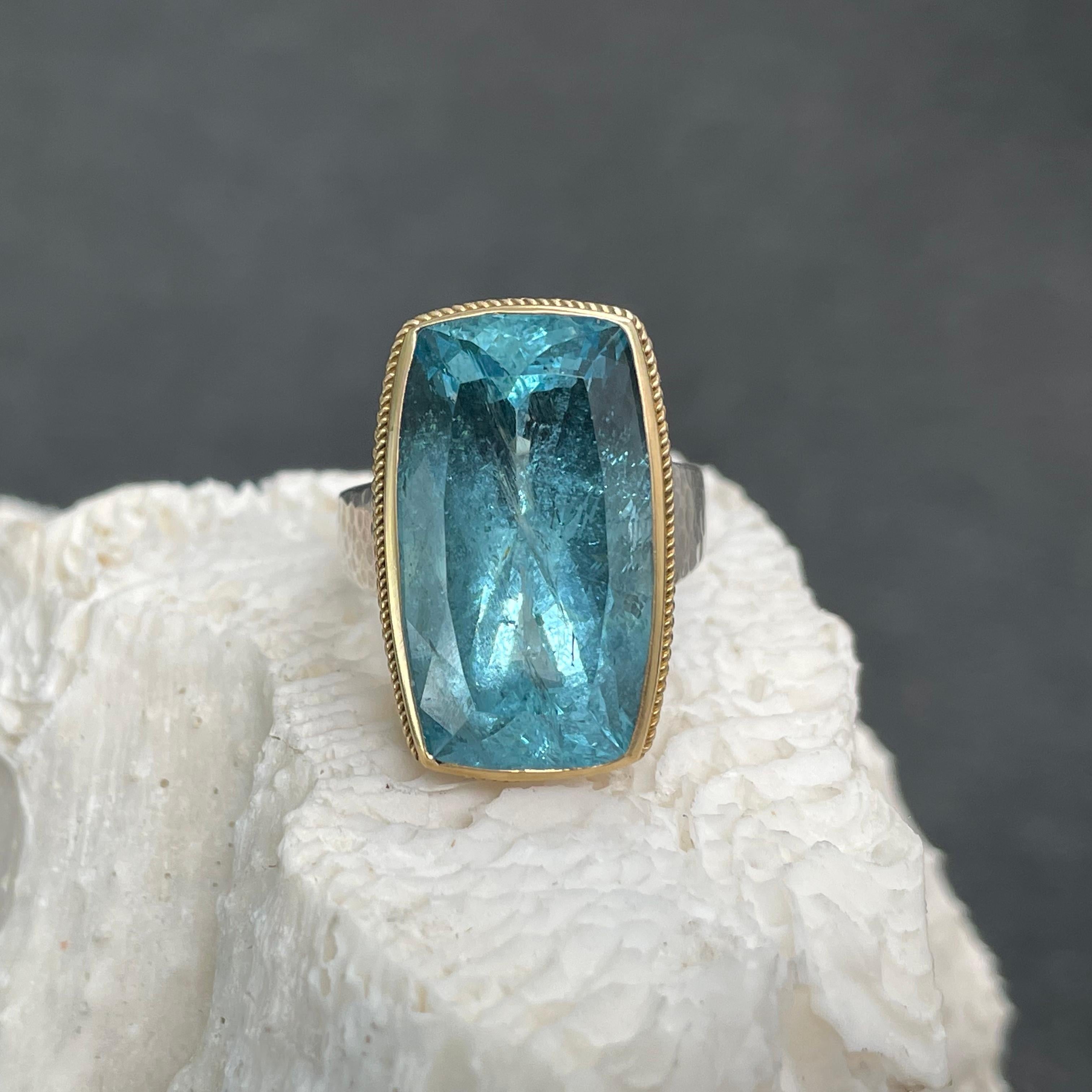 A deep color 10 x 14 mm long cushion shaped faceted Brazilian aquamarine is held in an 18K gold  