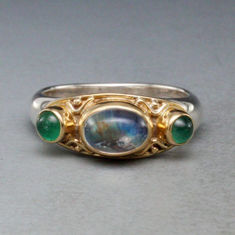 A nice combination of an oval 6 x8 mm flashing rainbow moonstone flanked by two luminous round 4 mm Zambian emerald cabochons rests in handcrafted 18K gold bezels with decorative 18K wire work and 