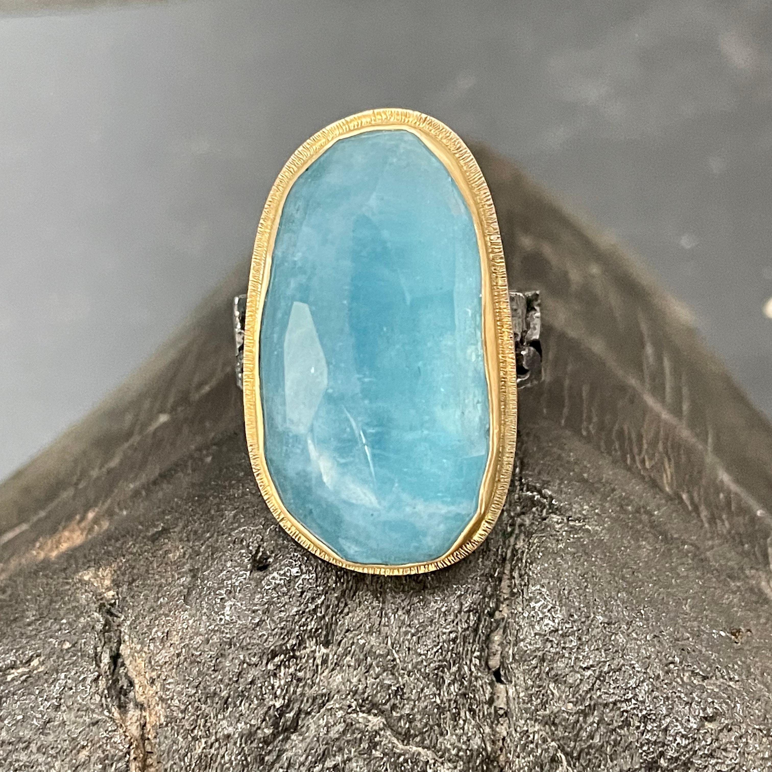A deep sea-blue irregular shaped 15 x 28 mm rose cut aquamarine is held in a slightly wavy 18K gold bezel surrounded by a line textured 18K highlight accent, all atop a wide organic textured oxidized sterling silver shank. The ring is currently