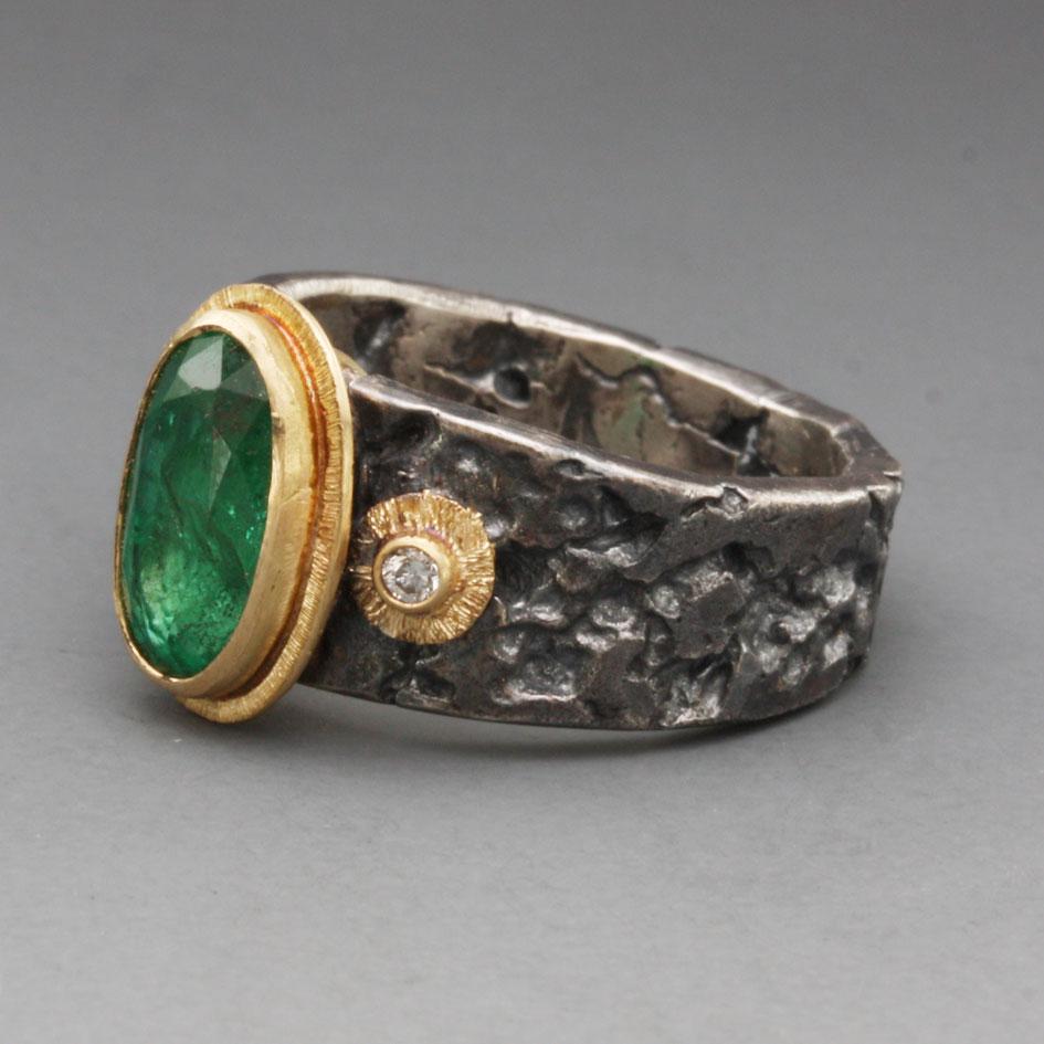 A lively 6 x 10 mm oval faceted Zambian emerald is set in an 18K gold bezel with a surrounding line textured gold double bezel. Two 1.8 mm VS1 diamonds are set to the sides with similar gold accompaniment.  All resting atop a comfortable flat and