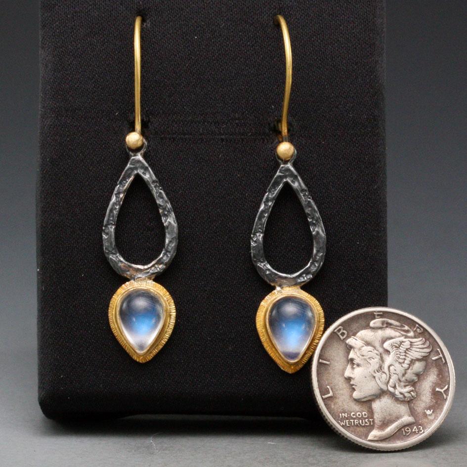 Two 6 x8 mm pear shaped rainbow moonstone cabochons surrounded by line texture 18K wide gold bezels form a complementary inverted pattern to the oxidized organic textured black silver components, all suspended below 18K gold safety clasp ear wires. 