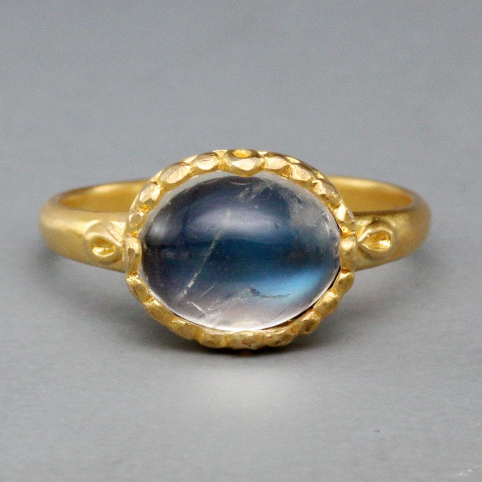 A shimmering 8 x 10 mm cabochon rainbow moonstone is set horizontally surrounded by delicately rounded flutes atop a slightly tapered shank with similar side accents.  All in matte-finish 18K gold.  This ring is currently sized 7.  It is easily