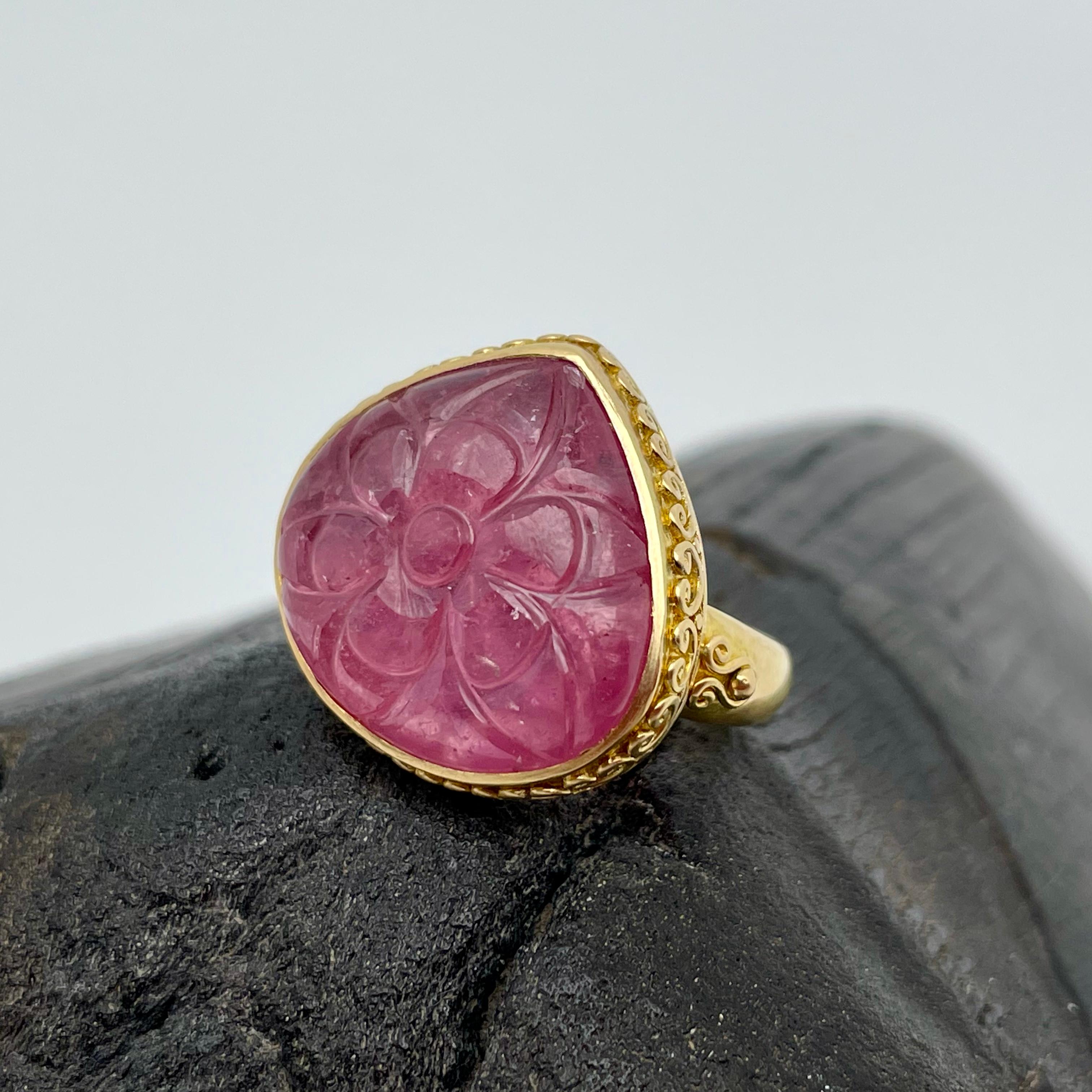 A translucent 17 x 19 mm drop shaped pink ruby cabochon is carved with a flower pattern and held in a handmade setting with delicate heart shaped wire accents on the bezel.  A tapered matte-finish shank with accents on the sides completes this