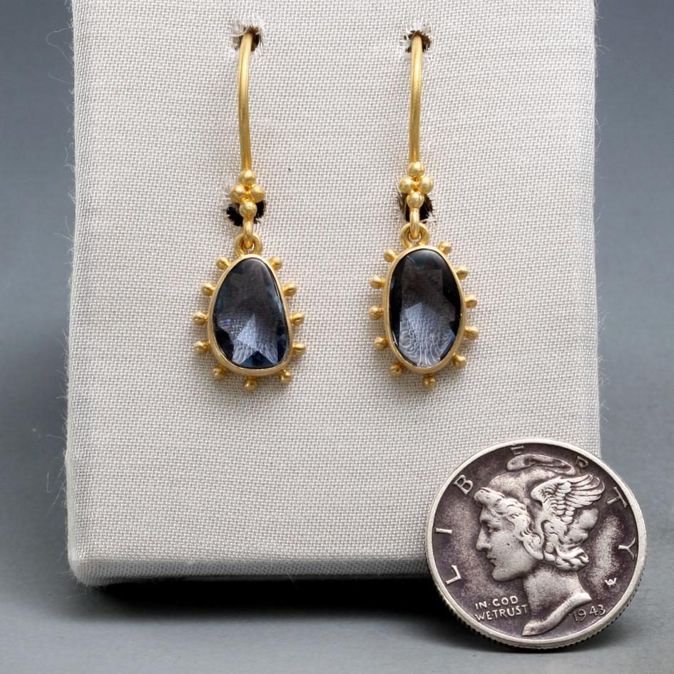 Two approximately 7 x 9 mm slightly irregular buff top blue sapphires are set in matt-finish 18K gold bezels surround by spaced 