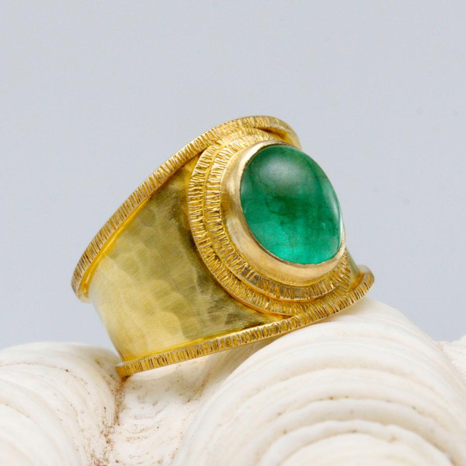 A limpid 7 x 10 mm oval Zambian emerald cabochon is highlighted by surrounding double line texture 18K gold bezels atop a comfortable wide hammered cigar band with similar line texture borders.  An elegant ancient-inspired handmade statement sure to
