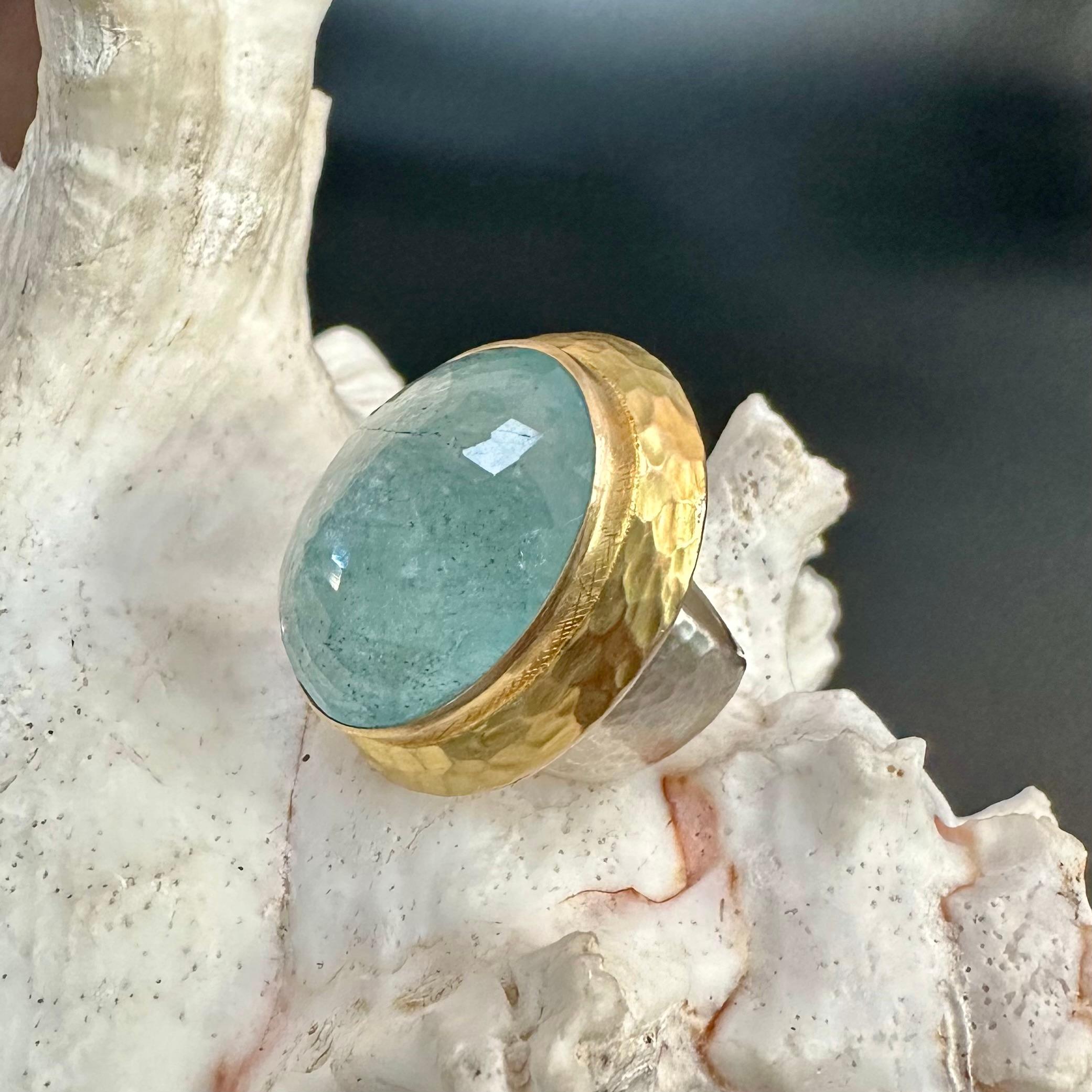 A sea-blue 18 x 25 mm oval rose faceted aquamarine is mounted longitudinally within a wide surrounding hammered double bezel 18K gold setting.  Along with the hammering, there is some cross-thatched motif along the base of the main bezel as well. 