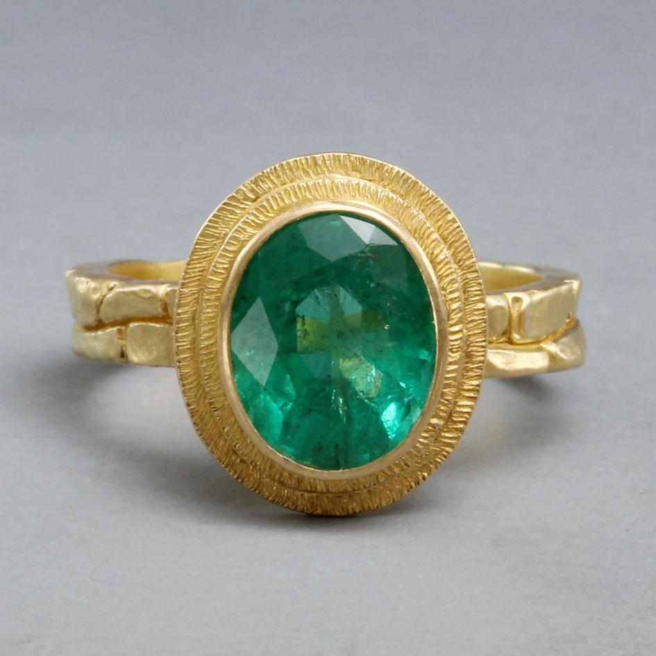 A crystalline high-grade 7 x 9 mm oval faceted Columbian emerald is the centerpiece of this one of a kind ring design.  The stone is set vertically surrounded by overlapping ridges of hand fabricated line-texture bezel atop a sculpted organic 18K
