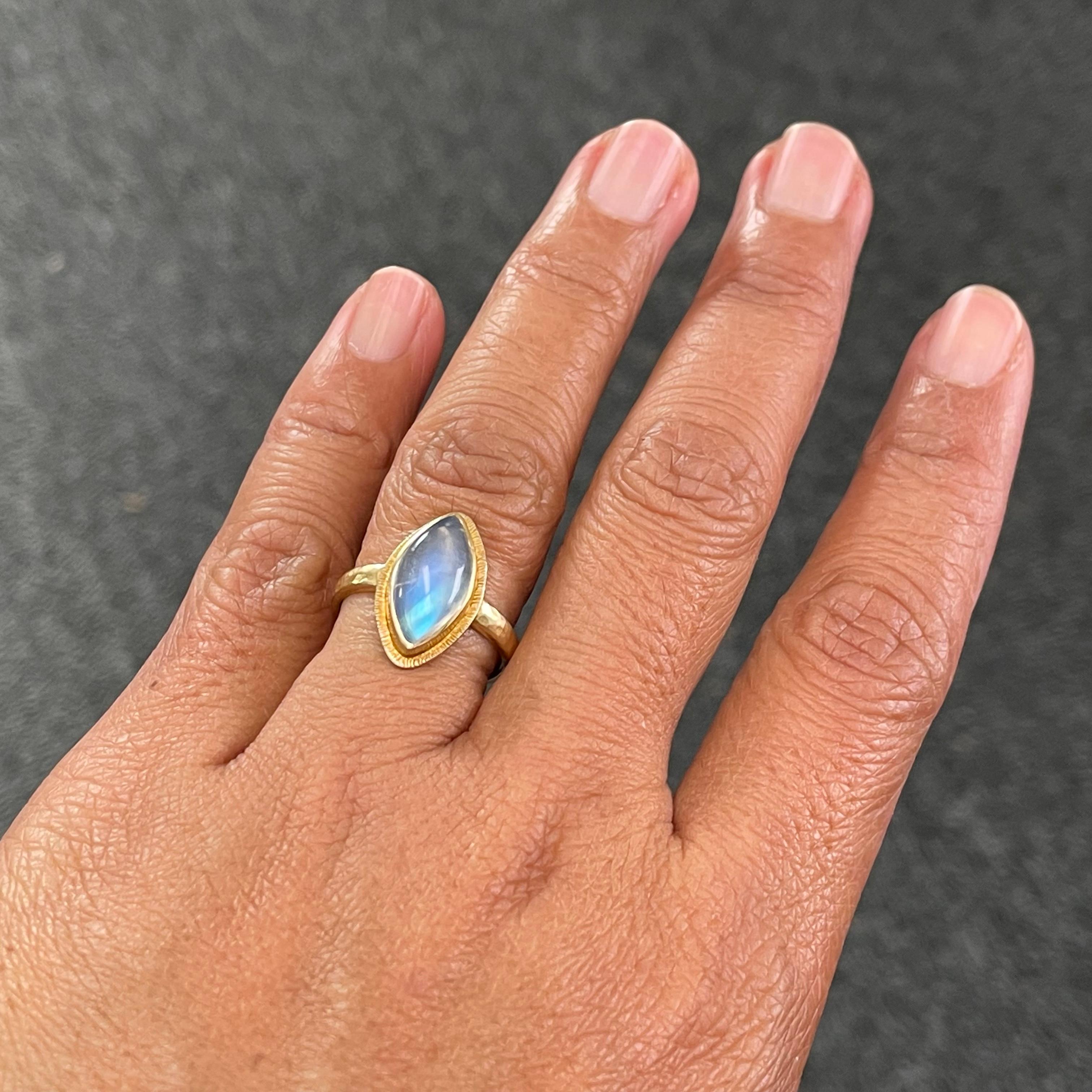 A lively 7 x 14 mm marquise shaped rainbow moonstone cabochon is held in a signature Steven Battelle line textured bezel atop a hammered matte-finish 18K shank.  The simple and organic look of the setting complements the shimmering blues of the