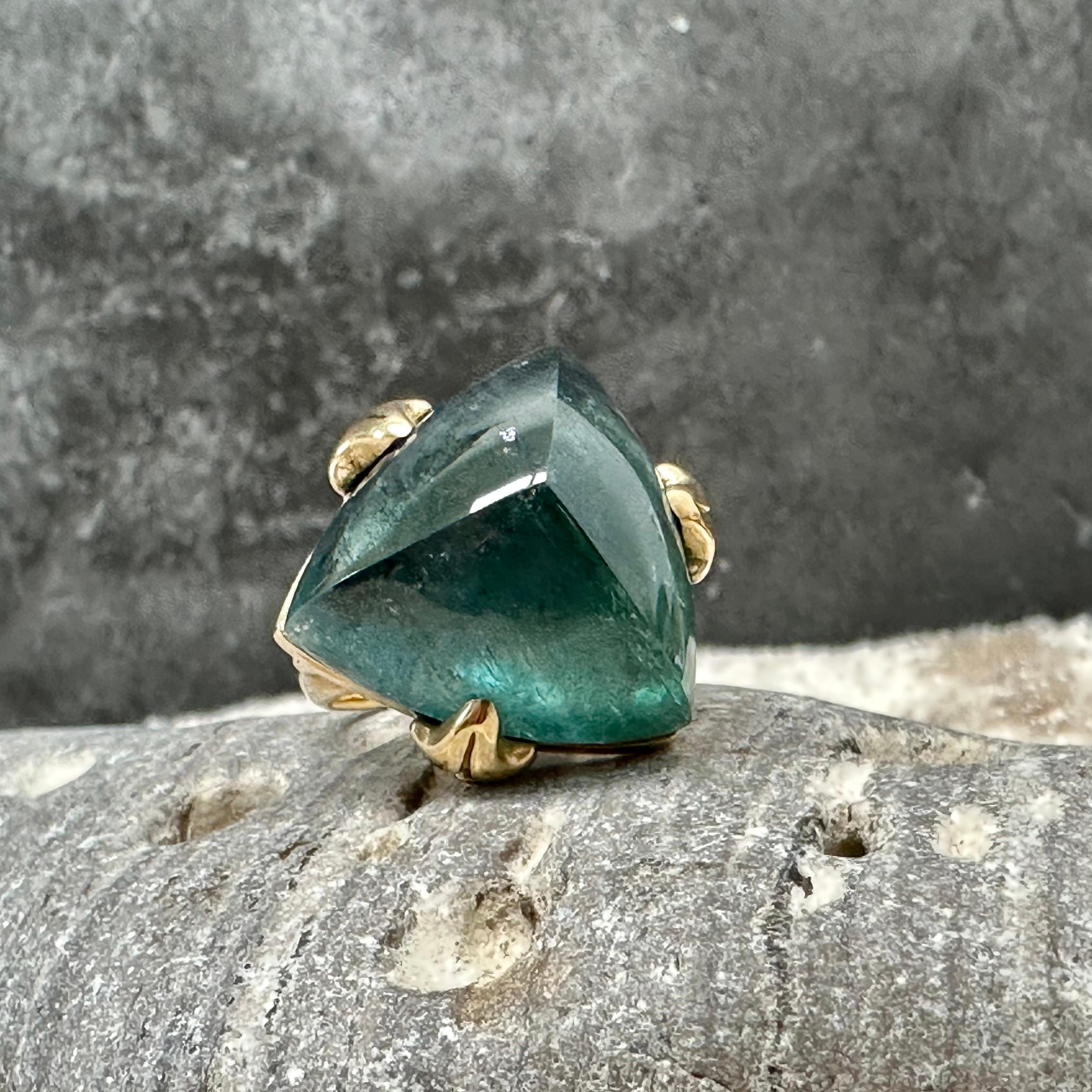 A 20 mm trillium cabochon of blue green Indicolite tourmaline is embraced by 3 carved prongs atop a tapered 18K shank attached  with a 