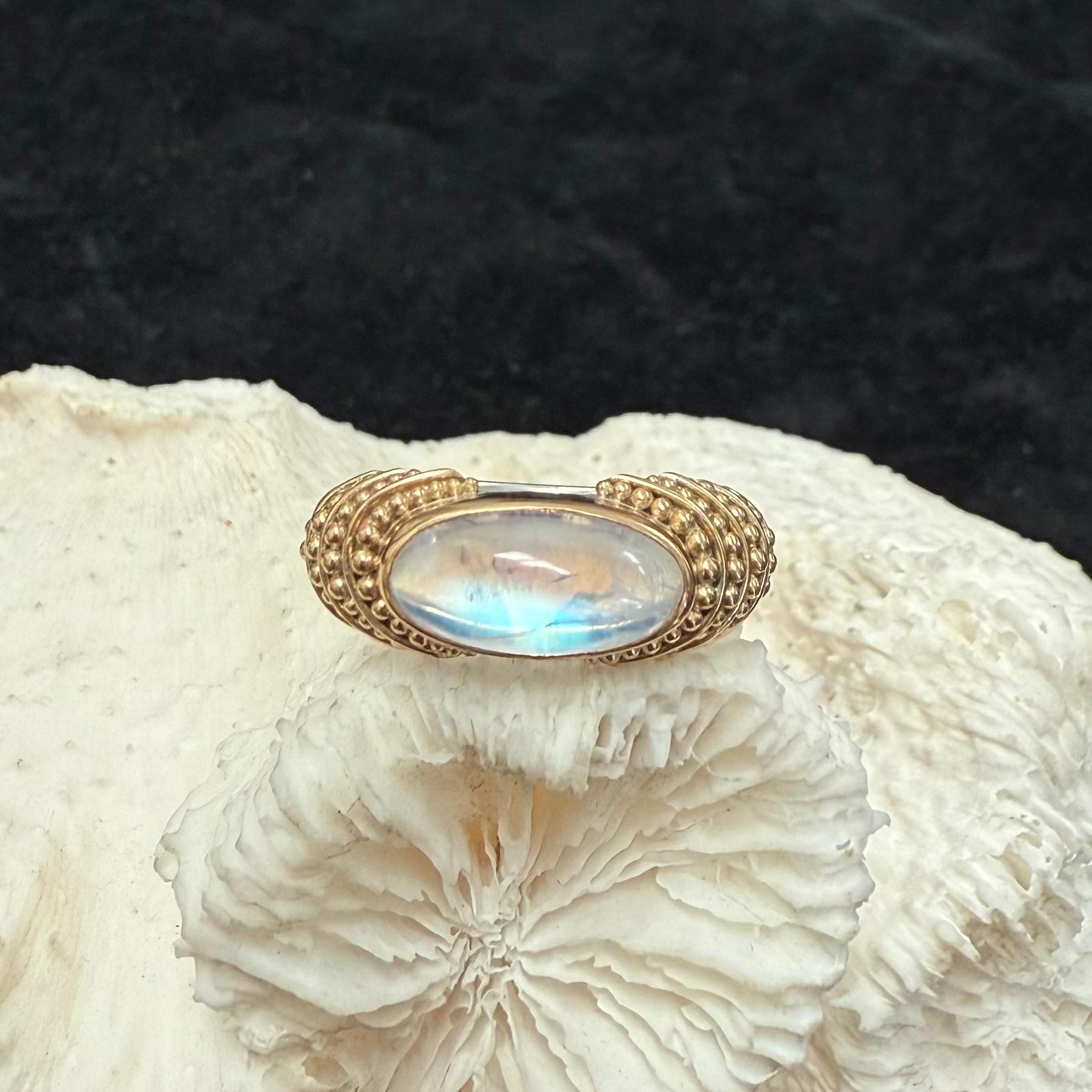 Una piedra lunar arco iris, larga y ovalada, de 6 x 12 mm, está engastada horizontalmente en un bisel de 22 quilates rodeado de capas escalonadas de granulado de 