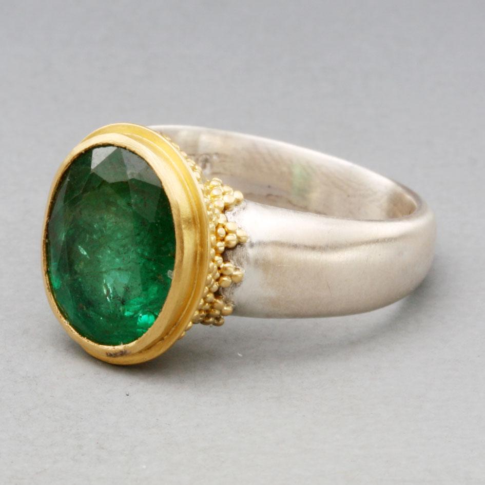 A lively 9 x 11 mm oval faceted Zambian emerald rests within a high-karat gold setting accented by handset geometric 