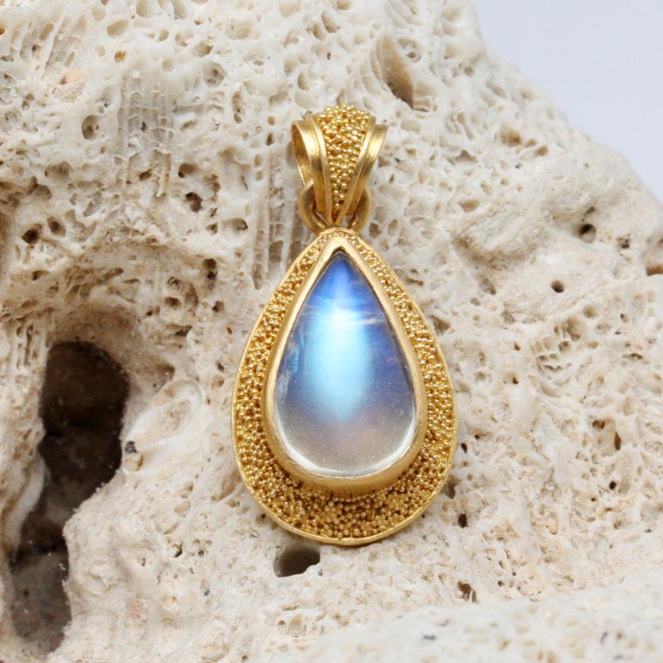 A shimmering blue 8 x 12 mm pear shaped rainbow moonstone is held in a high-Karat 22K gold bezel surrounded by delicate 