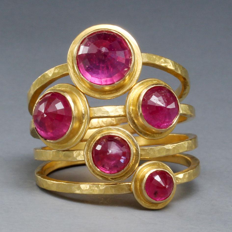 Five faceted round pink Thailand rubies ranging in size from 7 mm, 6 mm, 5 mm, 4 mm, and 3 mm are set reversed, culets up, with wide double bezel accents emphasizing the stones.  All are interspaced on 5 individual hammered matte-finish 18K gold