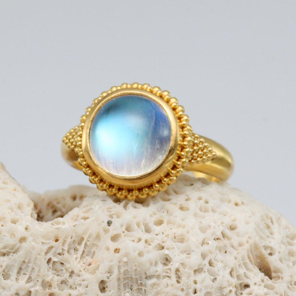 A glowing 10 mm rainbow moonstone cabochon is the centerpiece of this classic handmade geometrically granulated high Karat gold ring.  The stone is surrounded by a row of smaller atop larger granulation, and the substantial tapered shank has