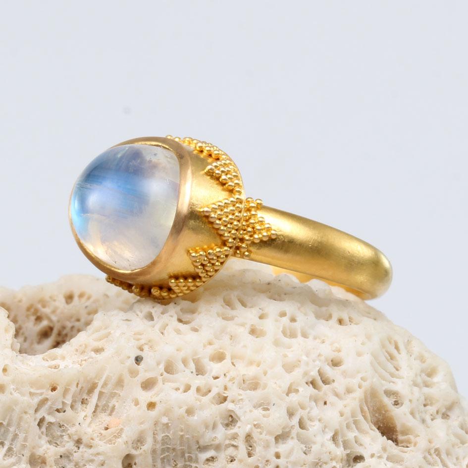 A lively blue flashing 9 x 11 mm rainbow moonstone is set horizontally in a signature cupped bezel granulated design ornament with meticulously hand placed tiny geometric 