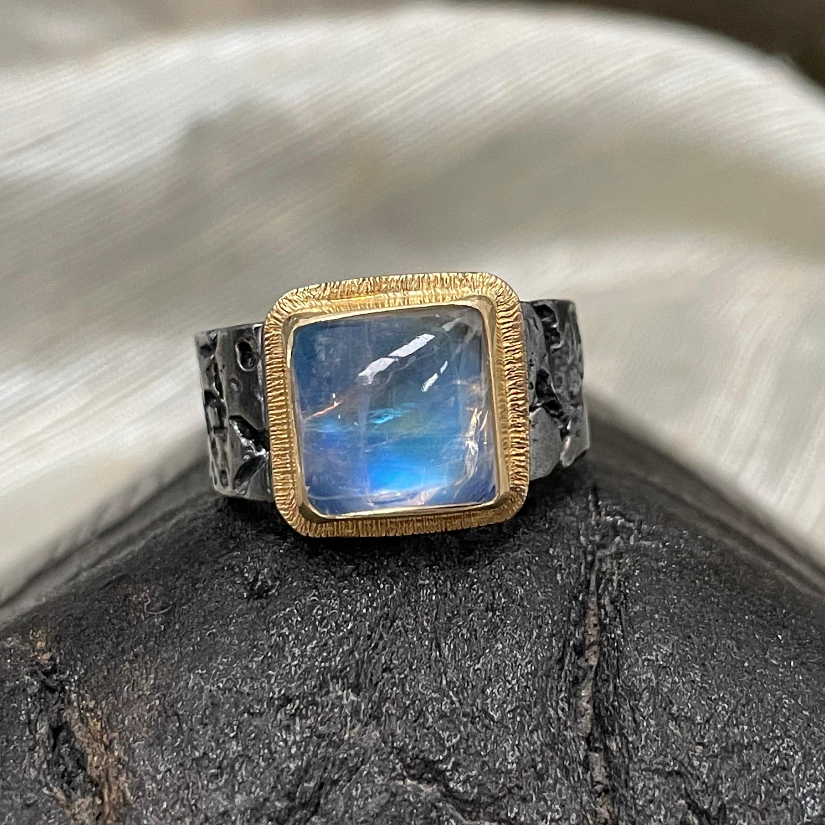 A beautiful 10mm square cushion rainbow moonstone is set atop an organic textured oxidized sterling slightly tapered flattish band in this juxtaposition of colors and textures.  The flashing blue of the moonstone contrasts beautifully with the