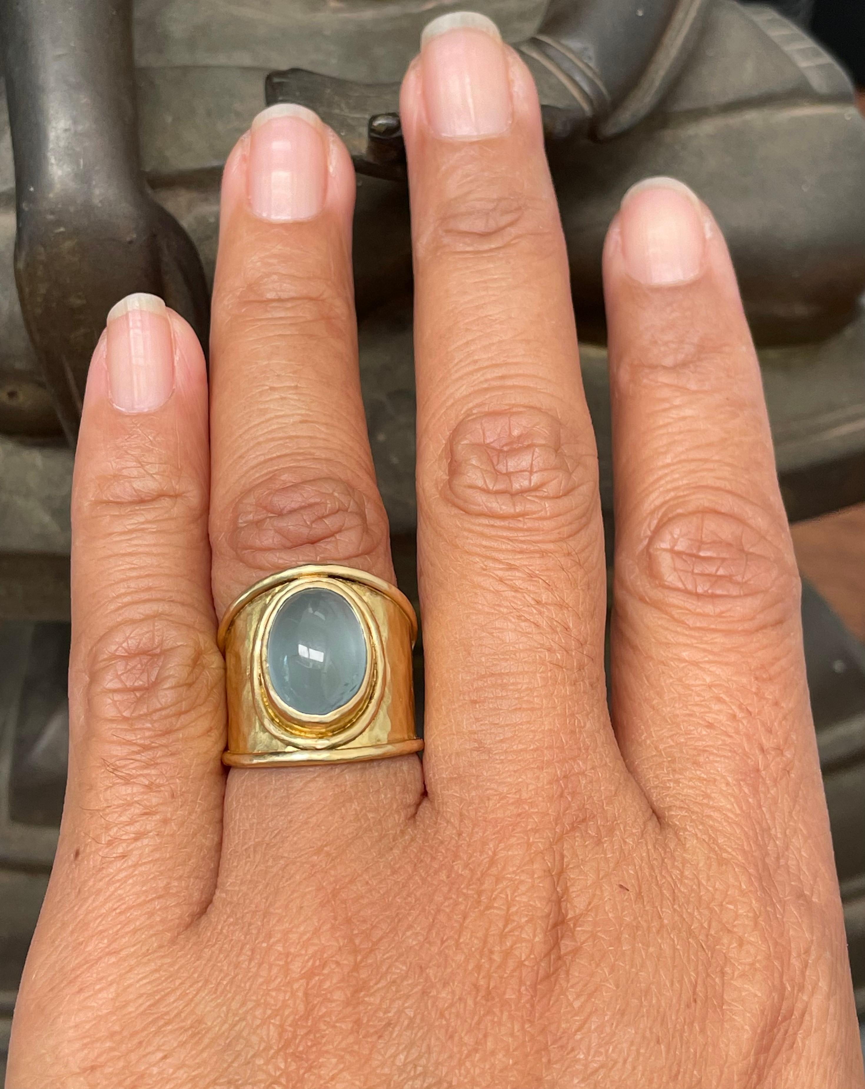 A nice sea green aquamarine 9 x 13 mm oval cabochon rests in a slightly cupped hammered 18K bezel surrounded by textured wire on a wide hammered textured 18K cigar band.  This stone has a lot of moonstone like chatoyancy.  This ring is sized 6.5.  