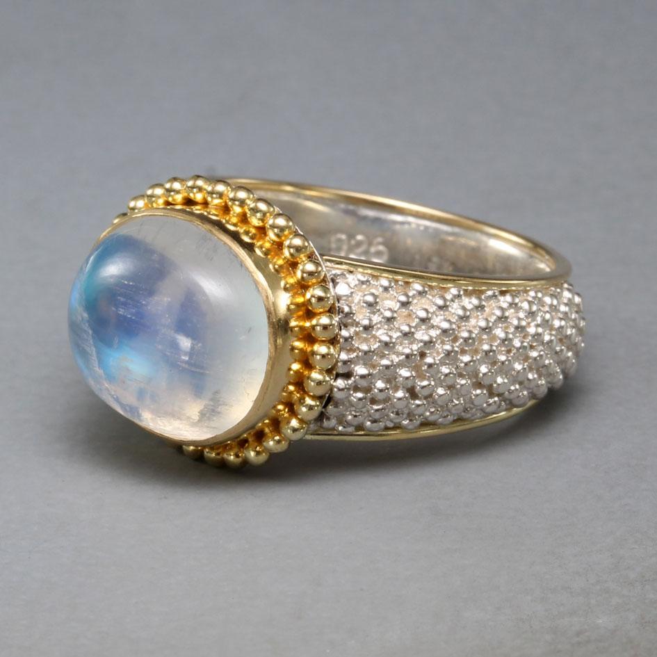 A blue flashing 10 x 12 mm rainbow moonstone cabochon is set horizontally in a 18K gold bezel surrounded by double row 