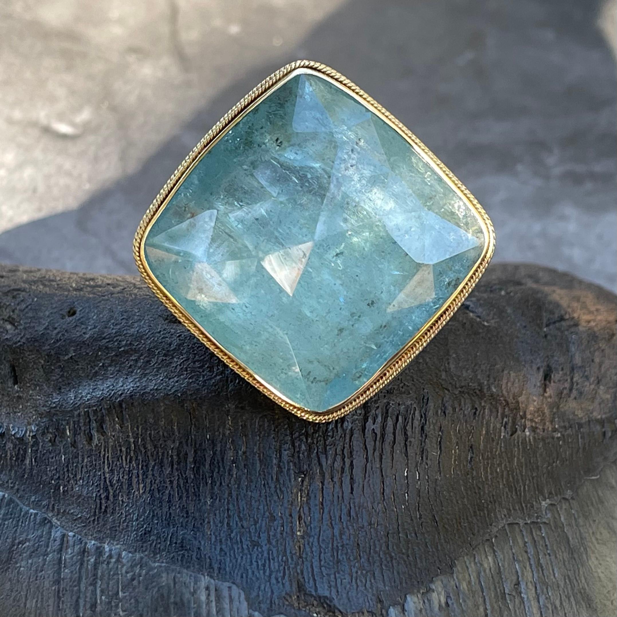 A large 27 mm x 27 mm light blue aquamarine cushion shaped faceted aquamarine is presented simply with an 18K gold bezel with double twist wire 18K accent in this Steven Battelle stunner. Mounted atop a sterling silver 