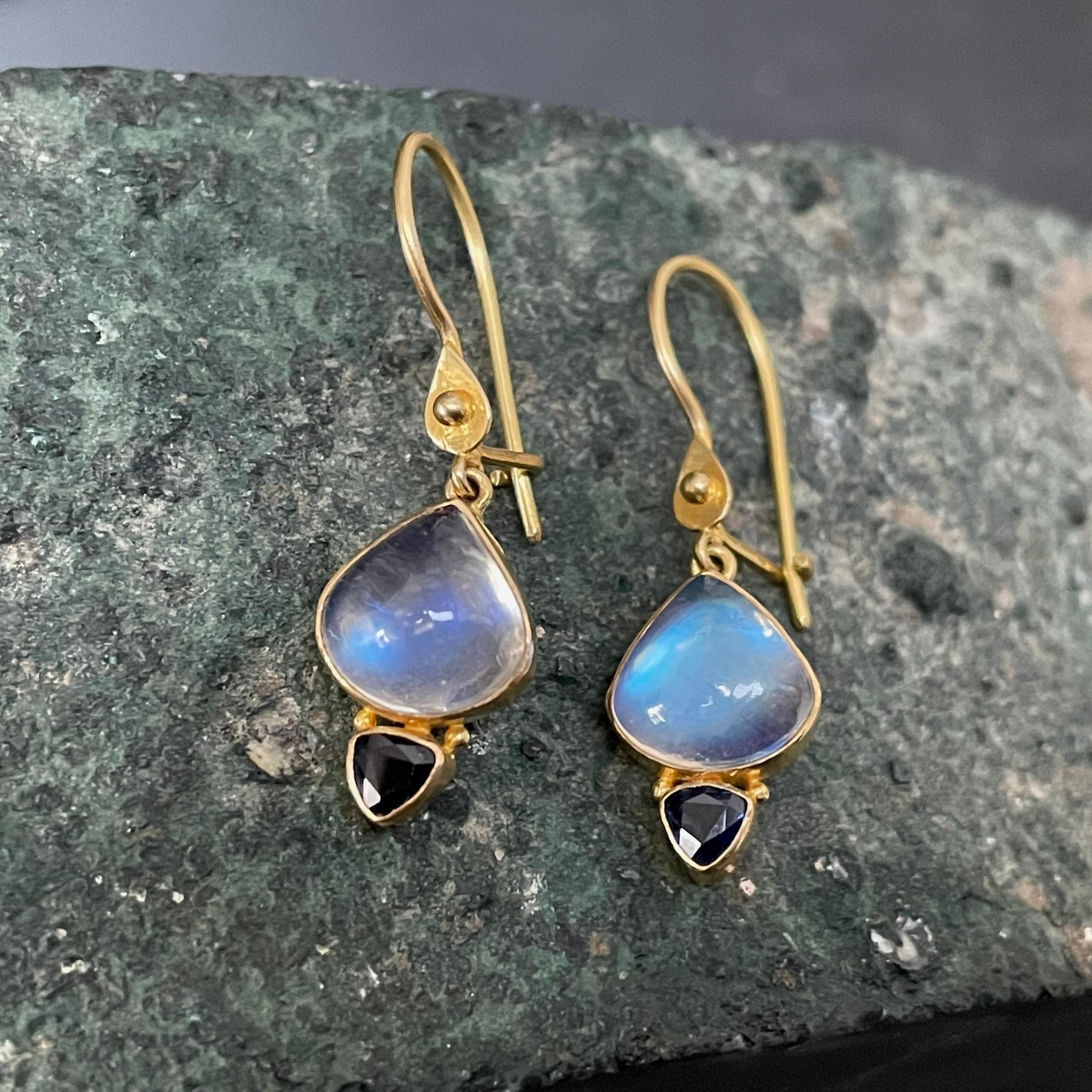 Two 3 mm trillium faceted blue sapphires are set in simple 18K bezels and attached below wide pear shaped 9 mm shimmering rainbow moonstones, all suspended below safety clasp wires with a single 