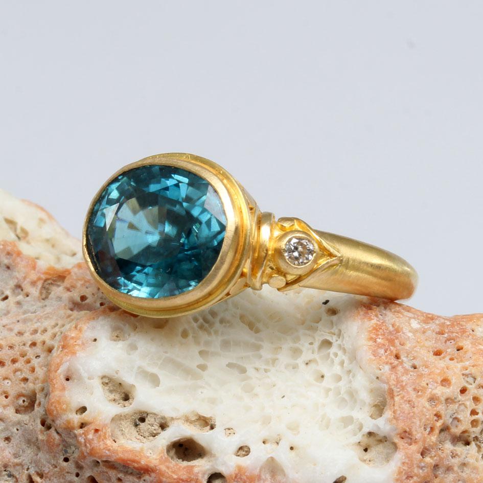 A highly lustrous, and bright  9 x 12 mm oval faceted Cambodian blue zircon is mounted horizontally in an artisanally crafted 