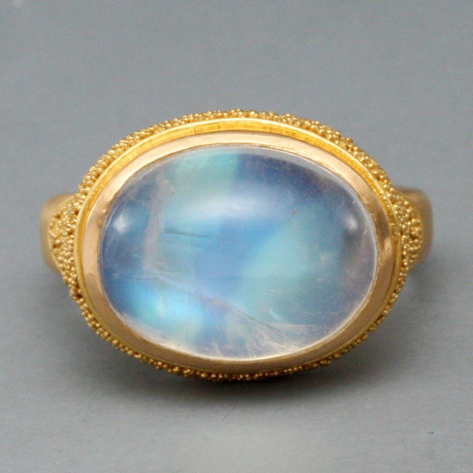 A blue flashing 10 x14 rainbow moonstone cabochon is set horizontally like a magical eye in a sightly cupped bezel surrounding by fine 