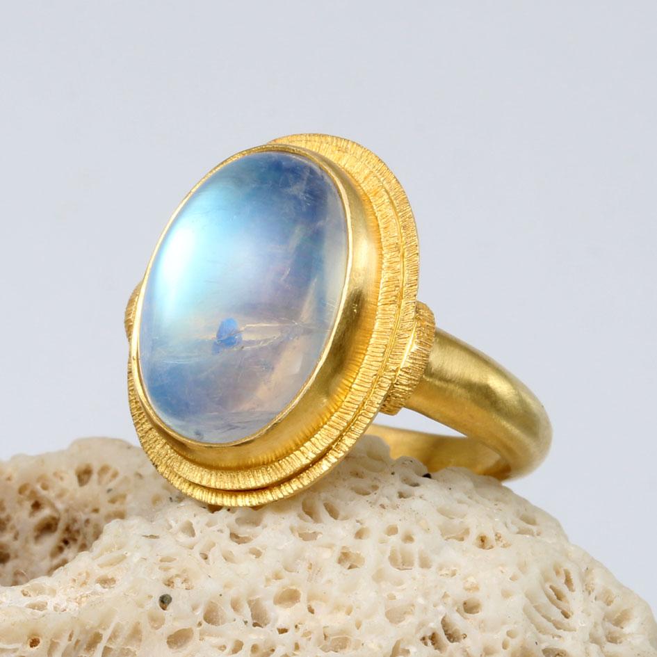 A very lively flashing blue 10 x 14 mm oval rainbow moonstone cabochon is prominently displayed surrounded by two concentric stepped organic line textured double bezels in this elegant 18K gold ring design.  The substantial matte-finish tapered